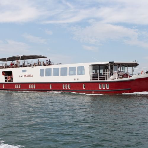 MS Ave Maria Flusskreuzfahrtschiff mit rotem Rumpf und weißem Aufbau auf ruhigem Wasser unter bewölktem Himmel. Passagiere auf Oberdeck sichtbar.