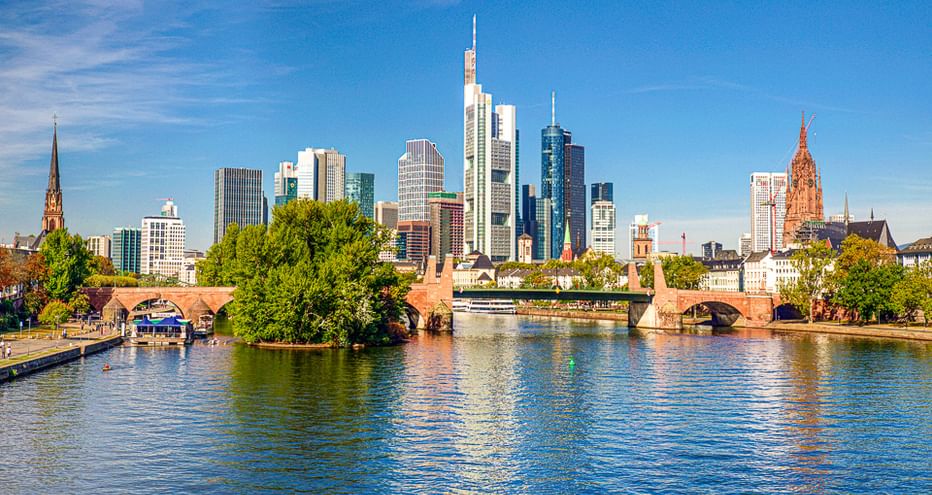 Frankfurter Skyline mit modernen Wolkenkratzern und historischen Kirchtürmen entlang des Mains. Steinbrücke überquert das Wasser unter blauem Himmel.
