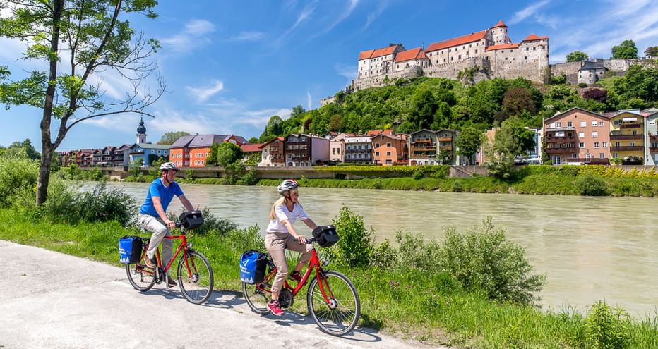 Zwei Radfahrer mit roten Rädern und blauen Taschen fahren auf einem Flussweg entlang der grünen Donau, mit einer Burg auf einem Hügel über einer Stadt.