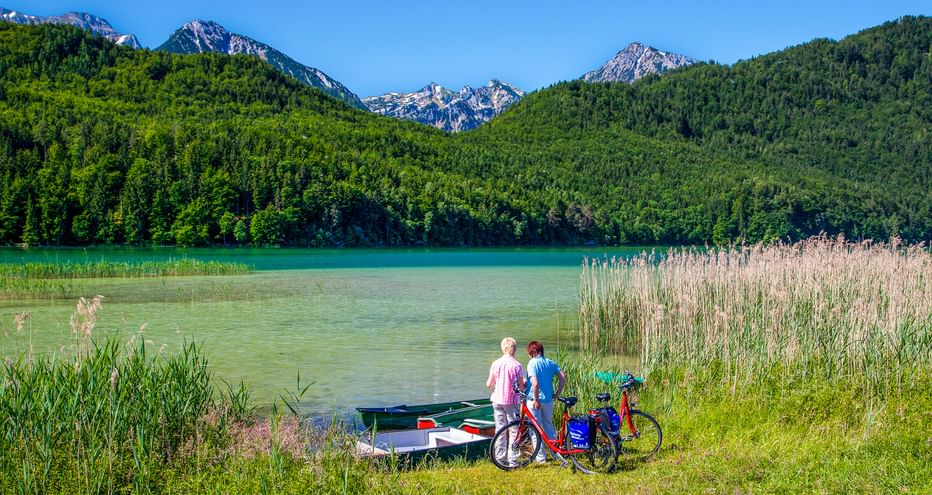 Zwei Radfahrer mit Rädern und Ruderboot am Ufer des Weißensees. Alpenberge und Wälder im Hintergrund unter blauem Himmel bei Füssen, Bayern.