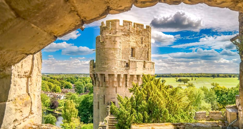 Blick auf den Steinturm von Warwick Castle durch einen verwitterten Bogen, mit grüner Landschaft und blauem Himmel dahinter.