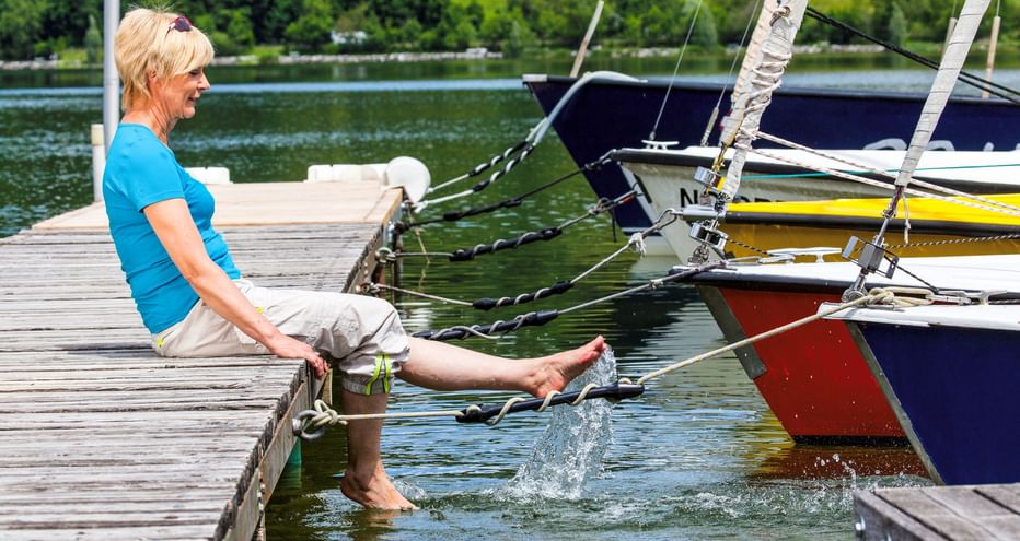 Frau in blauem Shirt sitzt auf Holzsteg mit Füßen im Wasser am Chiemsee. Bunte Segelboote liegen in der Nähe, grüne Hügel im Hintergrund.
