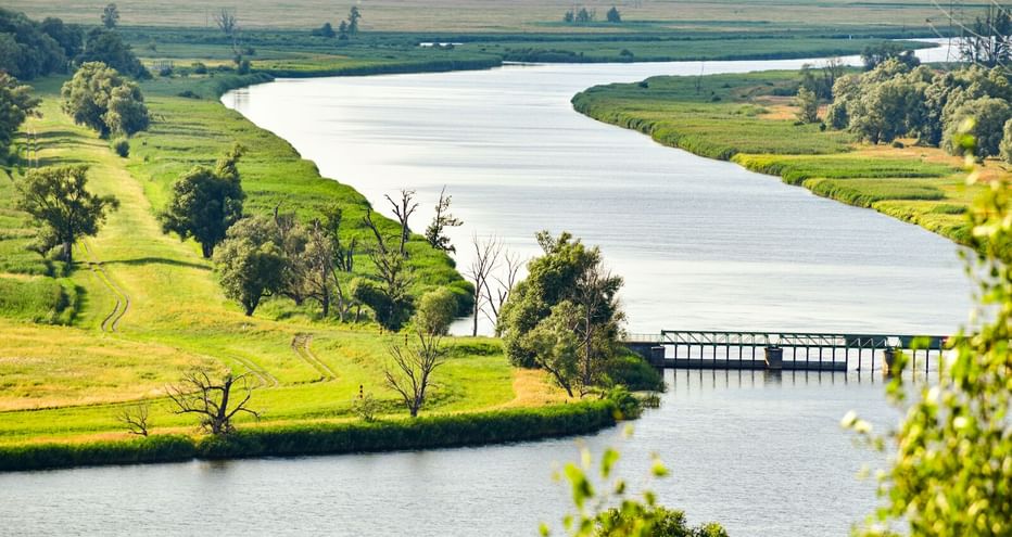 Aerial view of the Oder River winding through green meadows near Widuchowa. A small bridge crosses the river, with trees lining the banks.