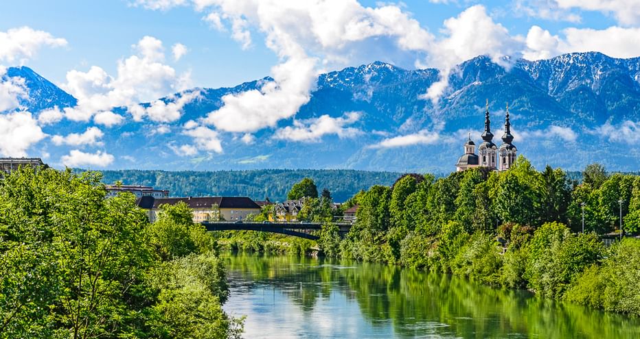 Fluss mit grünen Bäumen und Kirchtürmen, schneebedeckte Alpenberge im Hintergrund unter blauem Himmel mit weißen Wolken an der Alpe-Adria-Route.