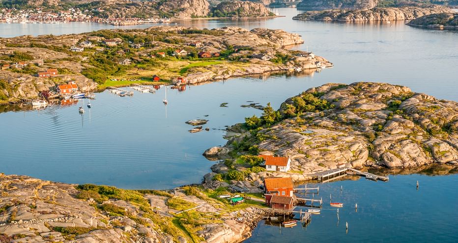 Luftaufnahme des schwedischen Westküsten-Schärengartens mit felsigen Inseln, roten Holzhäusern, Booten im ruhigen blauen Wasser und grüner Vegetation.