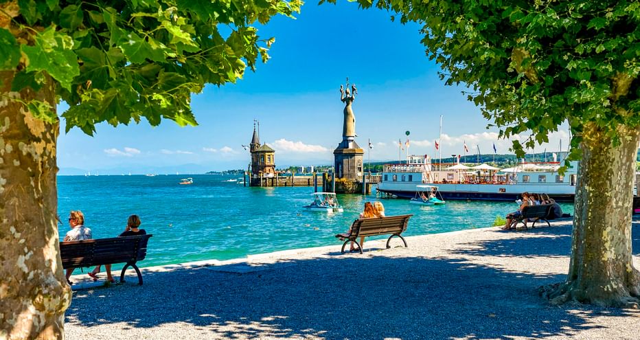 View through trees of Konstanz harbor on Lake Constance with the Imperia statue, lighthouse, and passenger ship. People sit on benches by the water.