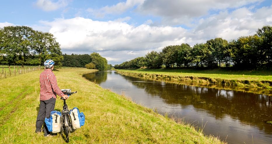 Radfahrer mit blauen Packtaschen steht am grasbewachsenen Flussufer neben der Ems. Grüne Wiesen und Bäume säumen beide Seiten unter bewölktem Himmel.
