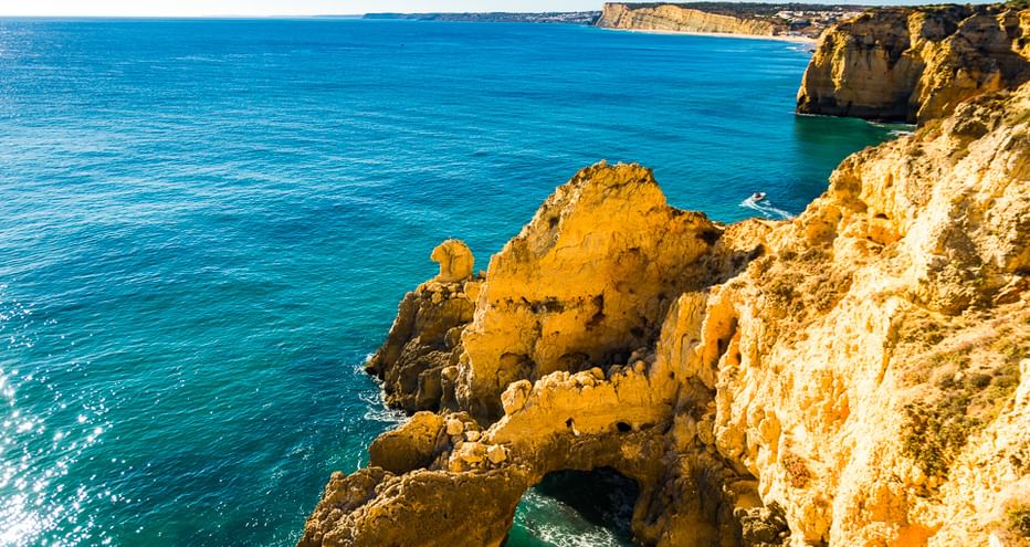 Goldene Sandsteinklippen ragen in das türkisfarbene Meer an der Algarve-Küste. Ein kleines Boot ist nahe den Felsformationen sichtbar.