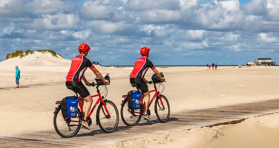 Zwei Radfahrer mit roten Helmen und Trikots fahren am breiten Sandstrand von St. Peter-Ording unter bewölktem Himmel. Sanddünen links sichtbar.
