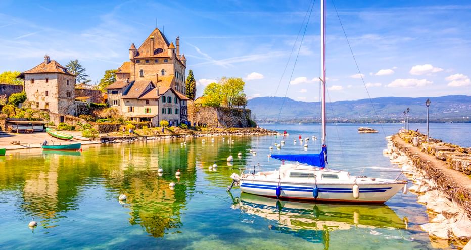 Blau-weißes Segelboot im klaren Hafen von Yvoire mit mittelalterlichen Steingebäuden und Kirche am Seeufer unter blauem Himmel.