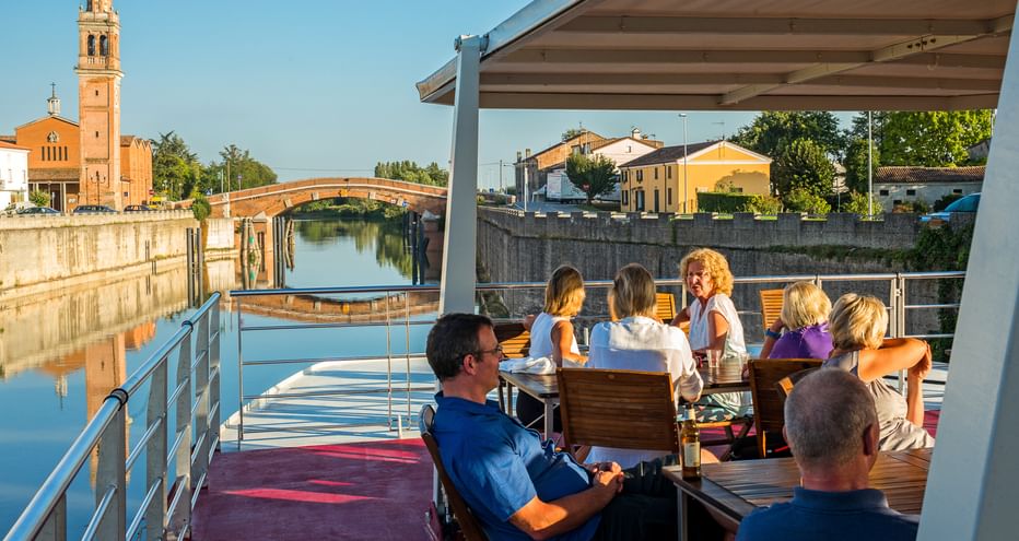 Passengers on the sun deck of MS Ave Maria in Portegrandi. View shows a canal with bridge, church tower, and Italian buildings under blue sky.