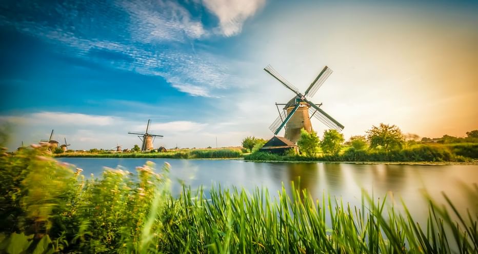 Traditionelle holländische Windmühle neben einem ruhigen Kanal mit grünem Schilf im Vordergrund. Blauer Himmel mit Schleierwolken.