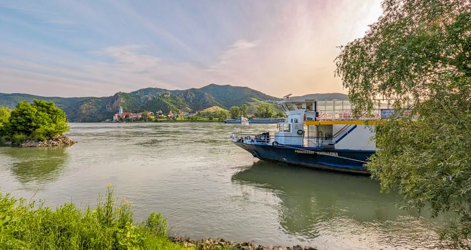 MS Prinzessin Katharina Flusskreuzfahrtschiff an der Donau mit grünen Ufern, Bergen und einem Dorf im Hintergrund.