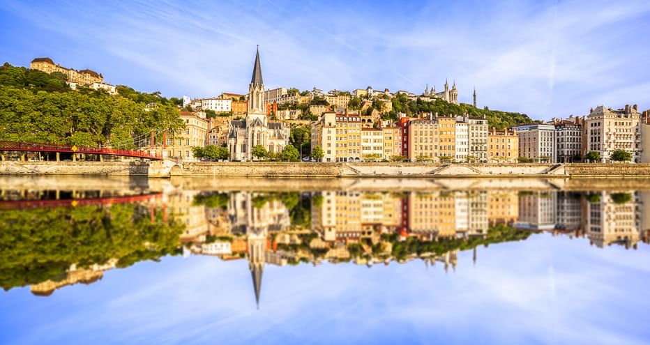 Historic Lyon riverfront with colorful buildings and church spire perfectly mirrored in the calm waters of the Rhône River under blue sky.