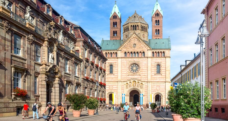 Radfahrer fahren über den Platz vor dem Speyerer Dom, einer romanischen Kirche mit Doppeltürmen und verzierter Fassade, eingerahmt von historischen Gebäuden.