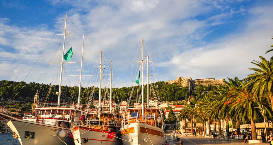 Drei hölzerne Segelboote mit hohen Masten liegen im Hafen von Hvar in Kroatien. Palmen säumen die Uferpromenade mit historischen Gebäuden am Hang.