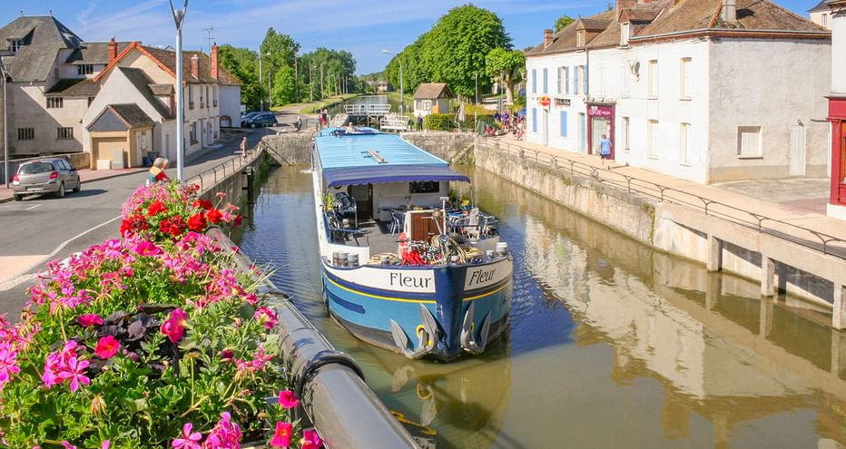 MS Fleur Flusskreuzfahrtschiff mit blauem Dach in einem schmalen Kanal in einem französischen Dorf, gesäumt von bunten Blumen und historischen Gebäuden.