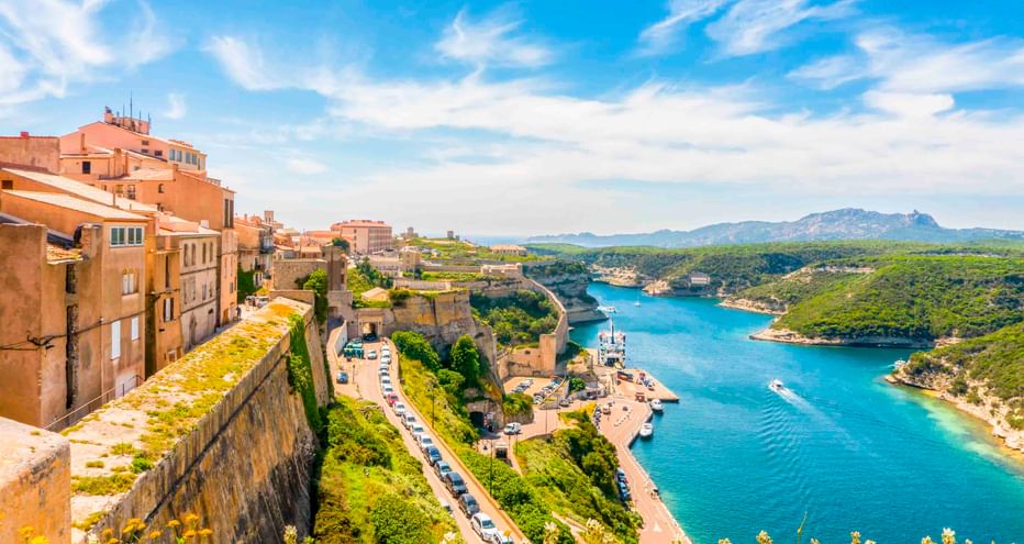 Panoramablick auf Calvi mit historischen Gebäuden auf einer Klippe über einer blauen Bucht mit Booten, grünen Hügeln und Bergen am Horizont.