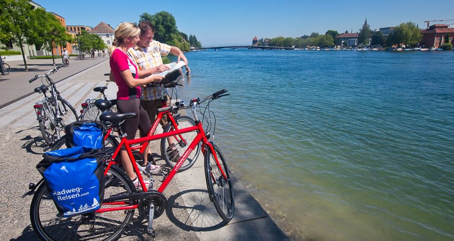 Paar mit Tourenrädern und blauen Packtaschen liest Karte an der Bodensee-Promenade. Klares blaues Wasser und Gebäude am gegenüberliegenden Ufer.