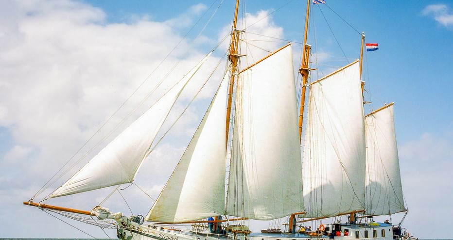 Traditionelles Segelschiff Elizabeth mit voll entfalteten weißen Segeln, segelnd auf grün-blauem Wasser unter teilweise bewölktem Himmel mit niederländischer Flagge.