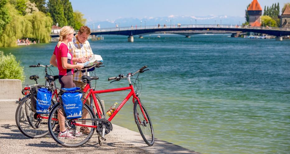 Zwei Radfahrer mit Tourenrädern lesen eine Karte am Seerhein in Konstanz. Brücke und historischer Turm über dem blauen Wasser sichtbar.