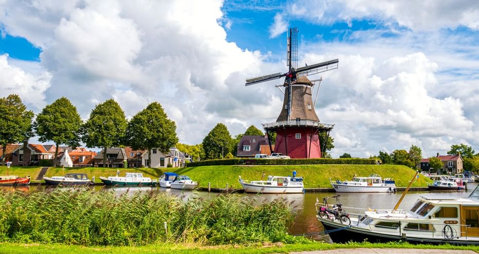 Traditionelle niederländische Windmühle auf Grasdamm in Dokkum mit Kanal, vertäuten Booten und Häusern unter blauem Himmel mit weißen Wolken.