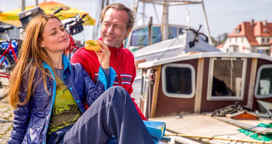 Lächelndes Paar in Radkleidung sitzt im Hafen der Holsteinischen Schweiz, Mann isst Sandwich. Boote und Gebäude im Hintergrund.