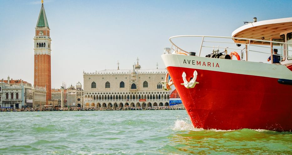 Rotweißes Schiff MS Ave Maria in der Lagune von Venedig mit Markusturm und Dogenpalast im Hintergrund unter blauem Himmel.