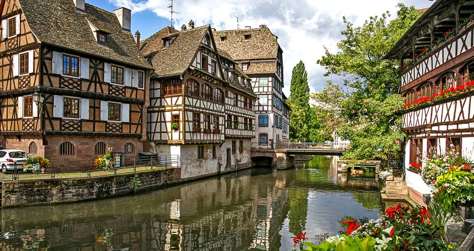 Traditionelle Fachwerkhäuser säumen einen ruhigen Kanal im Elsass. Eine Steinbrücke überquert das Wasser, bunte Blumen im Vordergrund.