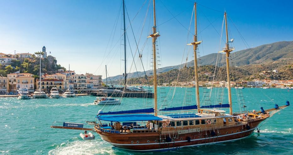 Hölzernes Segelschiff mit vier Masten in türkisfarbenem Hafenwasser. Griechische Küstenstadt mit weißen Gebäuden und Hügeln im Hintergrund.