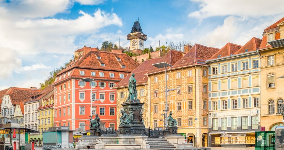 Hauptplatz square in Graz with ornate fountain monument in center, colorful historic buildings with red-tiled roofs, and iconic clock tower on hill.