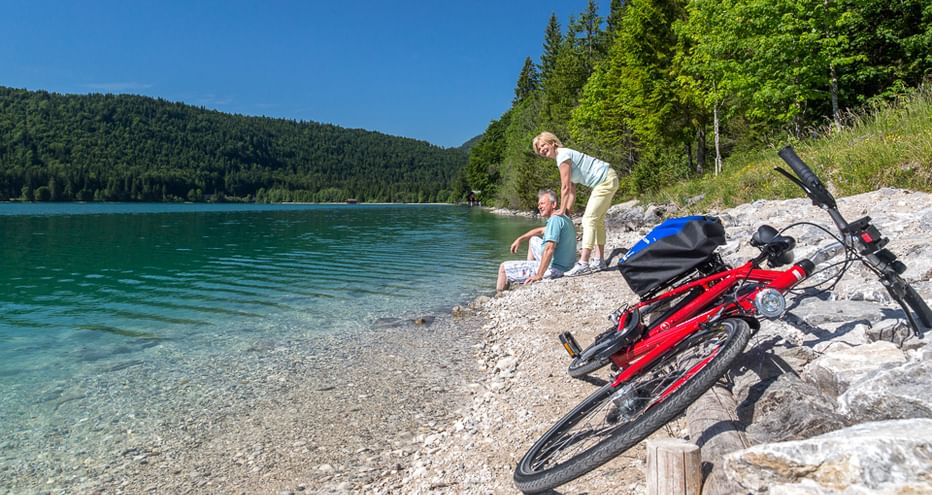 Zwei Radfahrer rasten an einem Kiesstrand neben einem türkisfarbenen See in Oberbayern. Zwei rote Fahrräder mit Taschen stehen im Vordergrund.