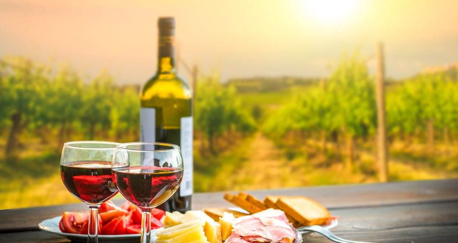 Zwei Gläser Rotwein mit Flasche, Käse, Prosciutto und Brot auf einem Tisch mit Blick auf einen sonnigen Weinberg in Italien.
