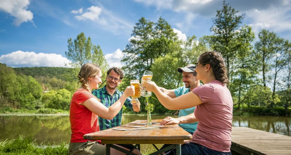 Vier Radfahrer stoßen mit Biergläsern an einem Holztisch an der Altmühl an, umgeben von grünen Bäumen unter blauem Himmel.