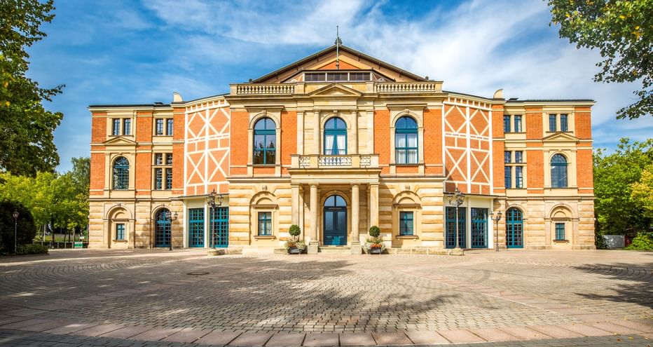 The Bayreuth Festspielhaus, a grand orange and cream-colored opera house with arched windows and decorative patterns, framed by green trees.
