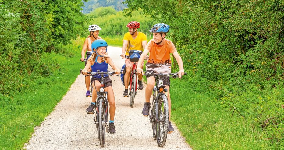 Vierköpfige Familie radelt auf Schotterweg durch grüne Landschaft. Zwei Kinder führen, gefolgt von zwei Erwachsenen, alle mit Helmen und bunter Kleidung.