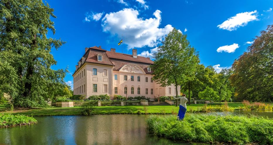 Schloss Branitz in Cottbus, ein beiges Barockgebäude mit rotem Dach, spiegelt sich in einem Teich. Üppige grüne Rasenflächen und Bäume umgeben das Schloss.