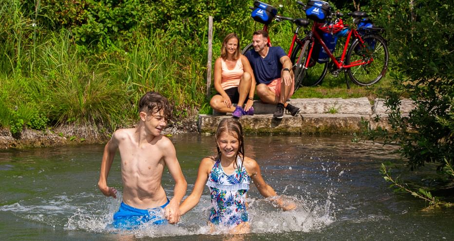Two children playing in a stream while parents sit on the bank with bicycles in the background at Lake Constance.