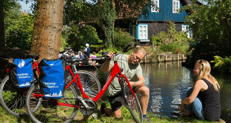 Zwei Radfahrer mit roten Rädern rasten an einem Kanal im Spreewald. Blaues traditionelles Haus am Wasser, umgeben von grünen Bäumen.