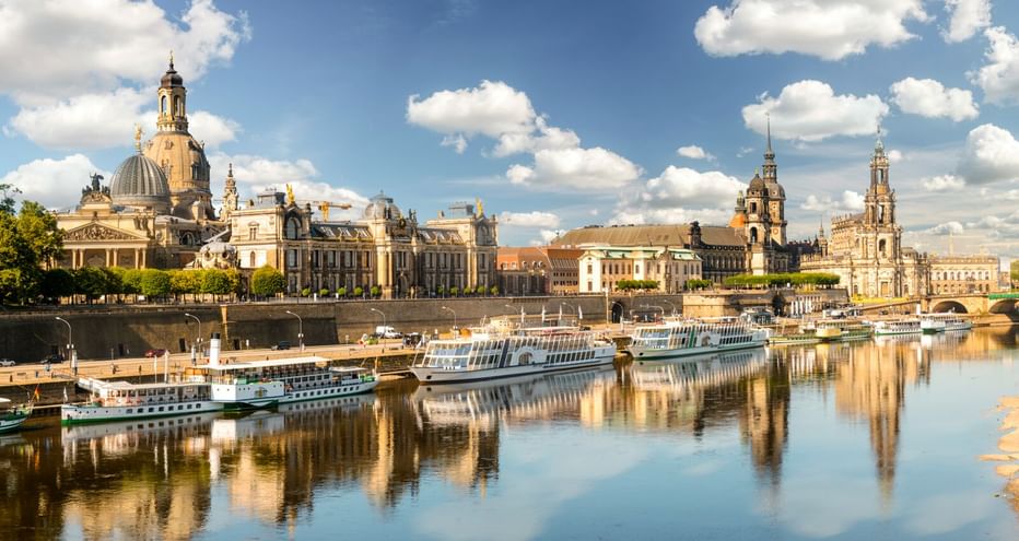 Dresdens historische Skyline entlang der Elbe mit barocker Architektur und Kuppelkirchen. Mehrere Flusskreuzfahrtschiffe liegen am Ufer.