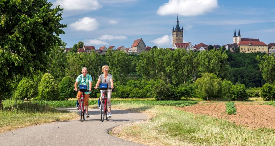 Two cyclists on a paved path with Bad Wimpfen's historic skyline in the background, featuring church towers and medieval buildings above green trees.