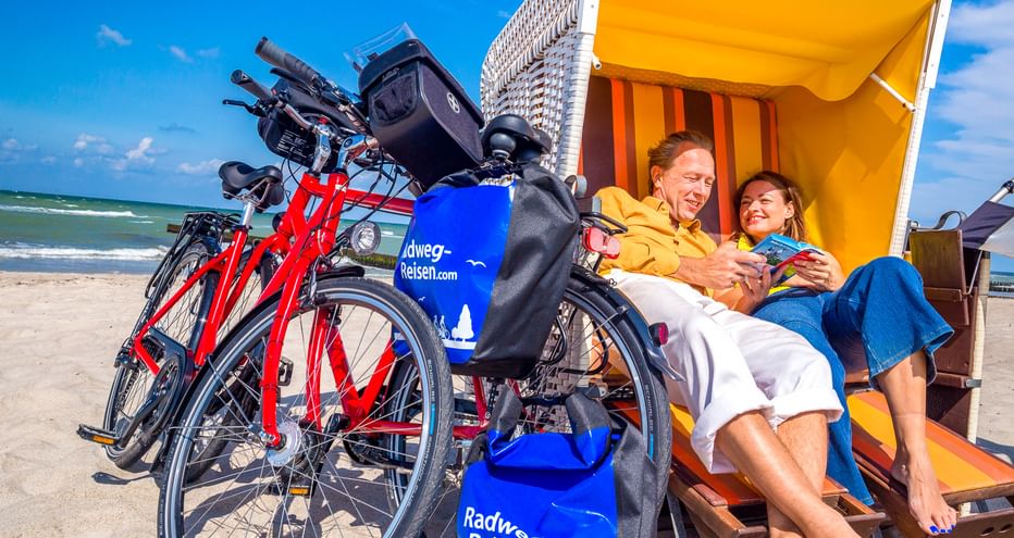 Zwei Radfahrer entspannen in einem gelb-orangen Strandkorb am Ostseestrand in Wustrow, mit roten Fahrrädern daneben unter blauem Himmel.