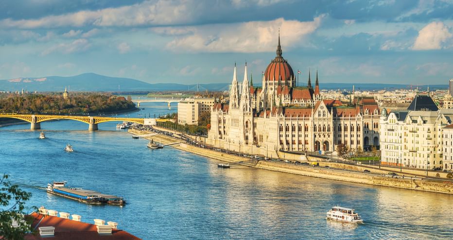 Budapester Parlamentsgebäude mit roter Kuppel am Donauufer. Blauer Fluss mit Booten, Brücke und fernen Bergen unter bewölktem Himmel.