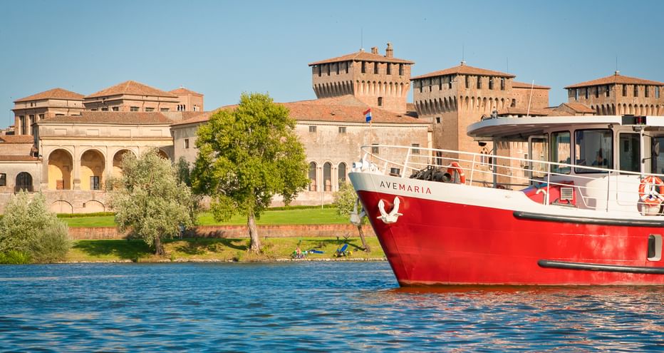 Red and white ship MS Ave Maria on blue water with historic Mantua fortress and towers in the background under clear sky.