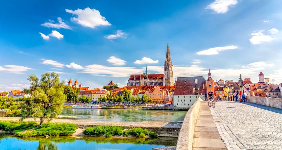 Historische Steinerne Brücke über die Donau zur Regensburger Altstadt mit Domturm und bunten mittelalterlichen Gebäuden unter blauem Himmel.