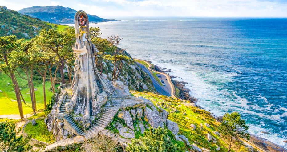 Virxe da Roca Steinmonument auf Küstenklippe in Baiona mit Blick auf den Atlantik, grüne Hänge und entfernte Berge.