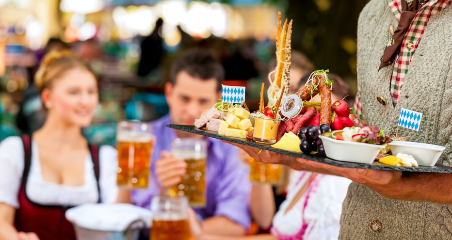 Kellner in traditioneller bayerischer Tracht hält Tablett mit Käse, Brezeln und Snacks. Gäste in Dirndl und Lederhosen mit Bier im Hintergrund.