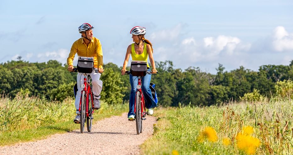 Zwei Radfahrer mit Helmen auf roten Rädern fahren auf einem Schotterweg durch grüne Wiesen mit gelben Blumen nahe Lillebælt.