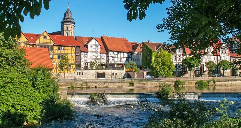 Fachwerkhäuser mit roten Dächern und Kirchturm an der Weser. Ein Wehr erzeugt fließendes Wasser im Vordergrund, eingerahmt von grünen Bäumen.