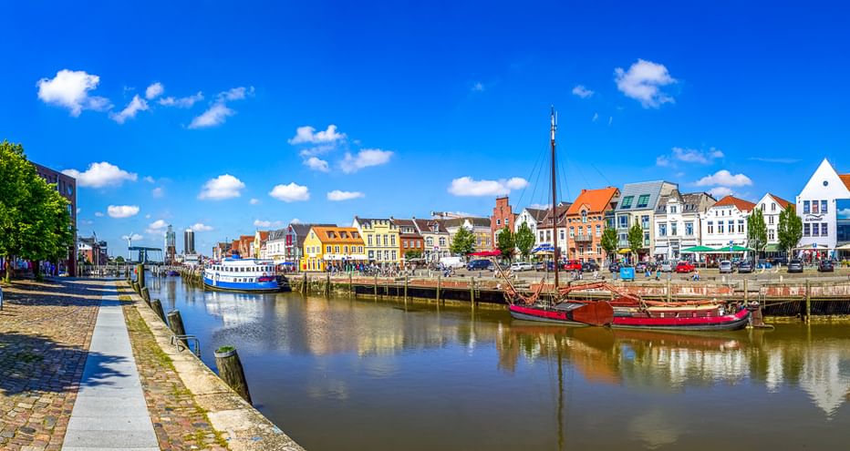 Historischer Hafen von Husum mit bunten Häusern entlang der Uferpromenade. Rote und blaue Boote liegen am Kai unter blauem Himmel.